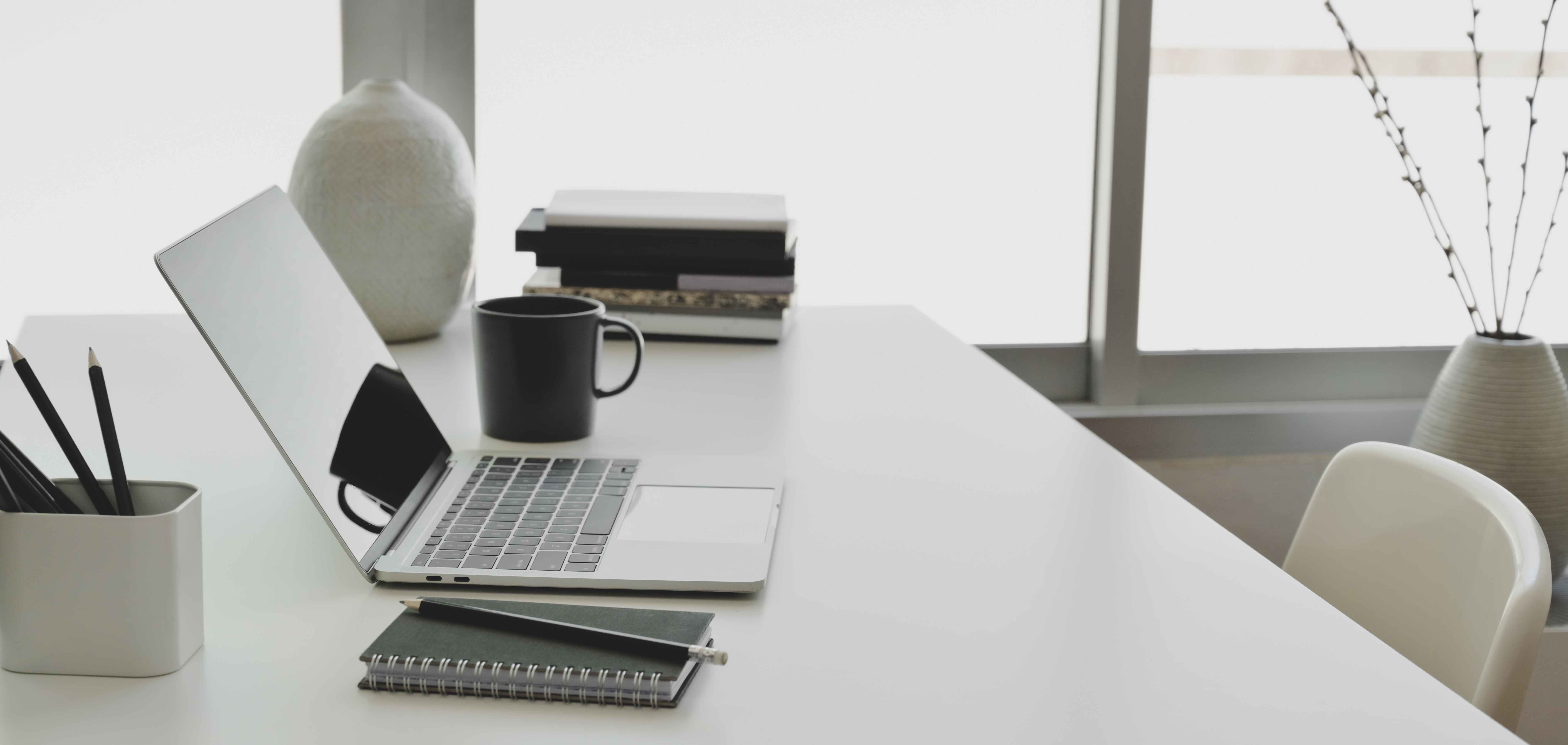 Comfortable office room with laptop computer and office supplies on white wooden table near the windows